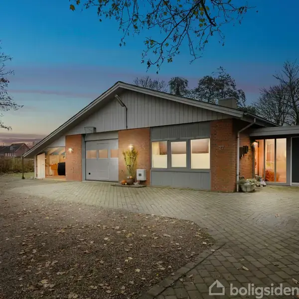 Et murstenshus med grå garageporte fremtræder oplyst ved skumringstid. Omgivet af en flisebelagt indkørsel og træer ses i baggrunden. Indendørs lys skinner gennem vinduerne.