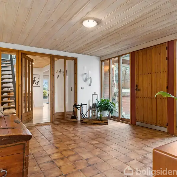 Entrérum med trægulvplanker og store vinduer, der lukker lys ind. Til venstre et kommode og spejl, i midten en trappe med glasparti, til højre en stor plante.