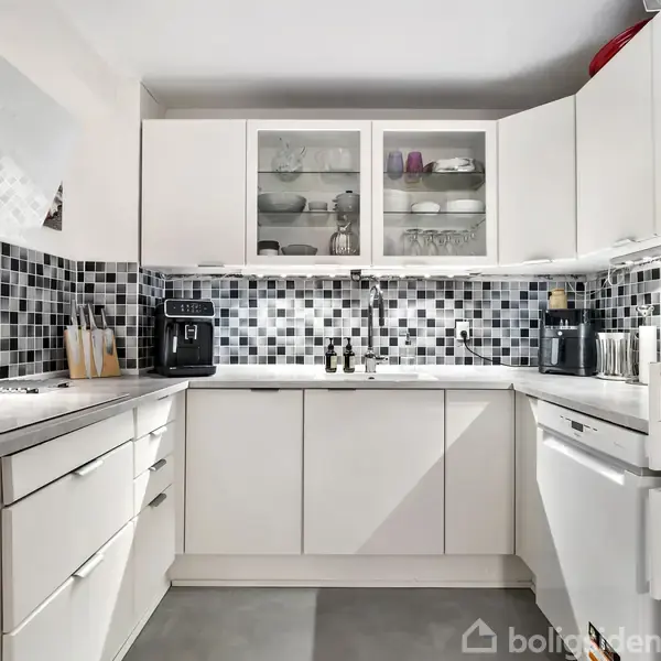 Hvidt køkken med grå mosaikfliser på væggene. En kaffemaskine står til venstre, omgivet af hvide skabe og skuffer. Et køleskab står til højre.