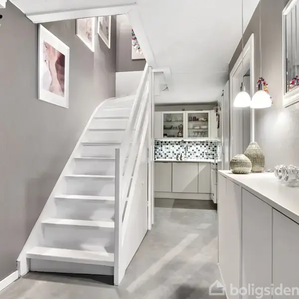 Trappe fører opad i moderne, lyst hjemmemiljø med grå vægge og hvid indretning. Hængende billeder og jakker til venstre; skab og planter til højre.