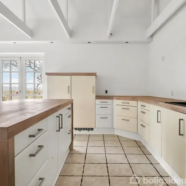 En tom stue med trætrappe til venstre fører opad. Store vinduer og terrassedøre giver udsigt til en have. Rummets vægge og loft er hvide med trægulve.