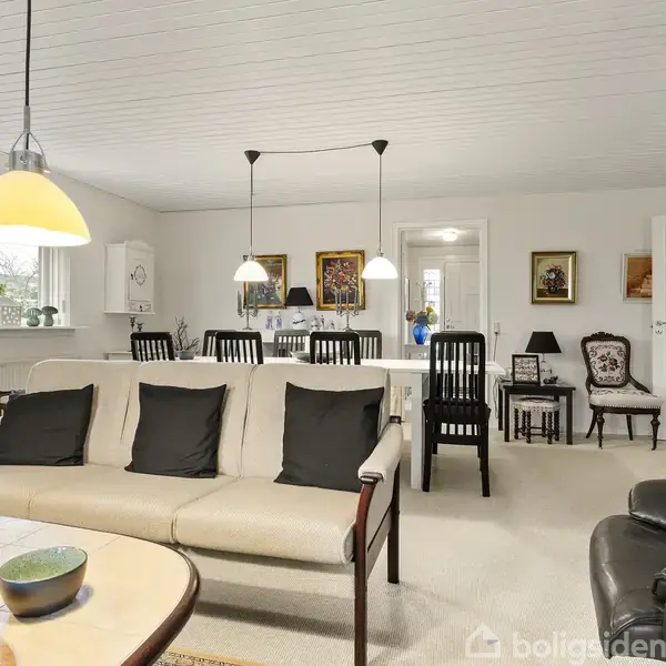 Sofa med sorte puder står i lys stue med spisebord og stole. Hængelamper lyser rummet op. Væggene er pyntet med billeder, og der er store vinduer. Ingen tekst fundet.