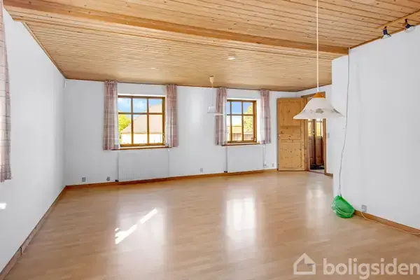 Et stort, tomt rum med trægulv og hvidmalede vægge. To vinduer med gardiner slipper lys ind. To lamper hænger fra loftet, og en grøn pose er på gulvet.