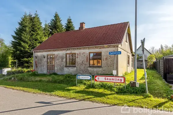 Et lille hus står stille i et landligt område. Et skilt ved siden af huset viser vej til "Skamling" og "Fjelstrupvej". Store træer danner baggrund.