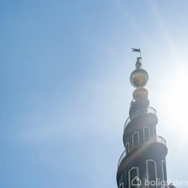 En spiralformet tårnspire med guldglobus og figur på toppen står mod en klar himmel. Sollys skinner skarpt fra højre side. Ingen tekst til stede.