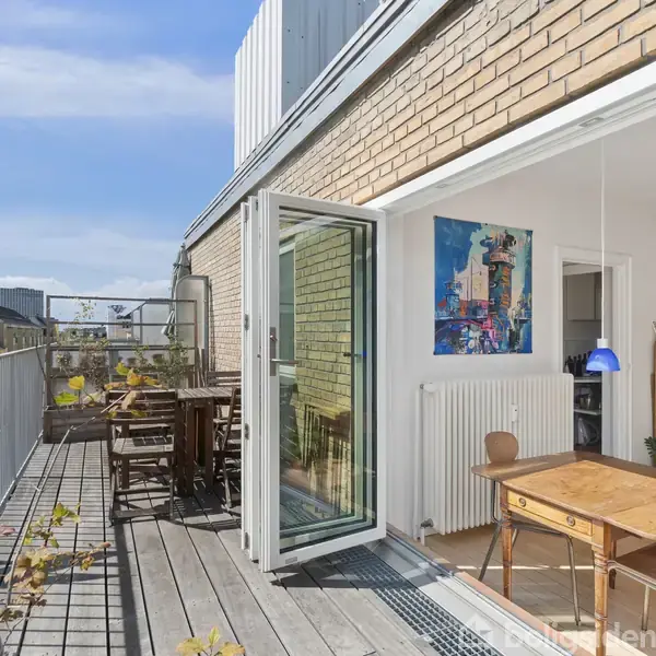 Altan med træplanker og bord-sæt under åben himmel. Døre fører ind til lys stue dekoreret med maleri på væggen og hængende lampe. Urban beliggenhed i baggrunden.
