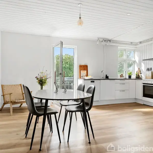 Et rundt spisebord med sorte stole står i et lyst køkken-alrum med åbne døre til en terrasse. Der er hvidt interiør og træmøbler på trægulve.