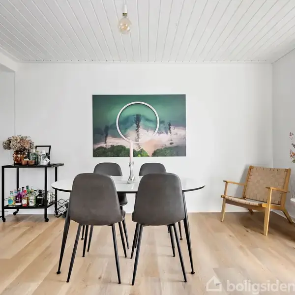 Spisebord med fire stole står i et lyst rum. En plakat hænger på væggen. Ved siden af står en lænestol og et bord med blomster på en lys trægulv.