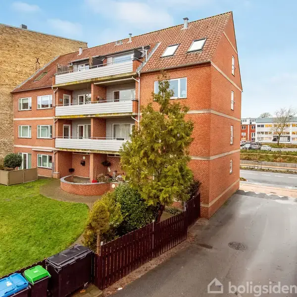 En murstensbygning med altaner står stille ved en grøn græsplæne. Omkringliggende huse ses ved siden af en asfalteret vej. Farverige skraldespande er placeret tæt ved muren.