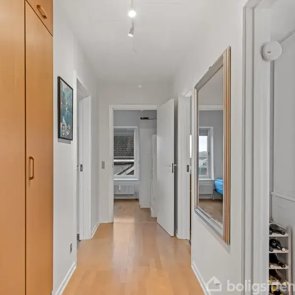 Gang med trægulv fører til et rum med blå og hvide elementer. Venstre væg har skabe og et maleri, mens højre væg har et spejl og vitrineskab.