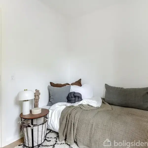 En seng dækket med puder og et tæppe i et minimalistisk værelse. Ved siden af står et lille bord med en lampe og dekorative genstande. Åben dør til andet rum.