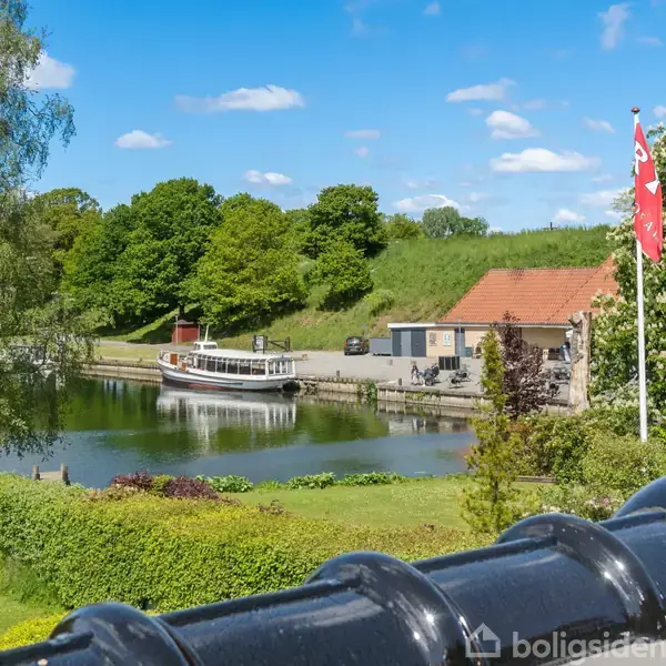 En båd ligger stille ved en lille havn, omgivet af frodig natur og en bygning med rødt tag. En dansk flagstang står nær ved, under en klar blå himmel.