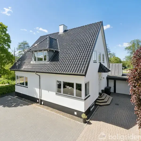 Et hvidt, to-etagers hus med sort tag står i en solrig have med en stor flisebelagt indkørsel. Omkring huset ses grønne træer og buske i et boligkvarter.