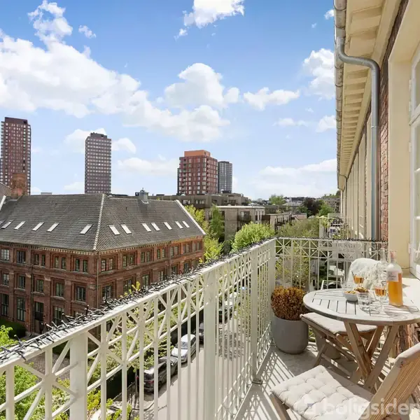 En balkon med et lille rundt bord og to stole indeholder drikkevarer. Den vender ud mod en udsigt over byens tage og højhuse under en blå himmel med skyer.