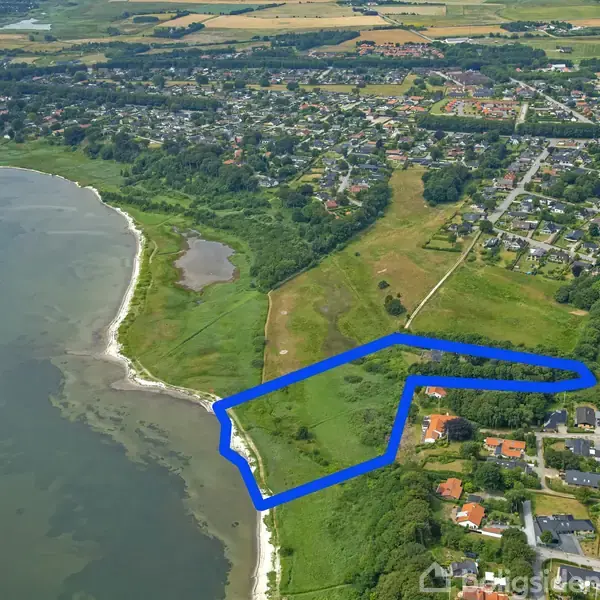 Parcel afgrænset med blå linje ved kysten. Området er grønt og frodigt, omgivet af huse og marker. Kystlinjen grænser op til en rolig vandkant. Byområde i baggrunden.