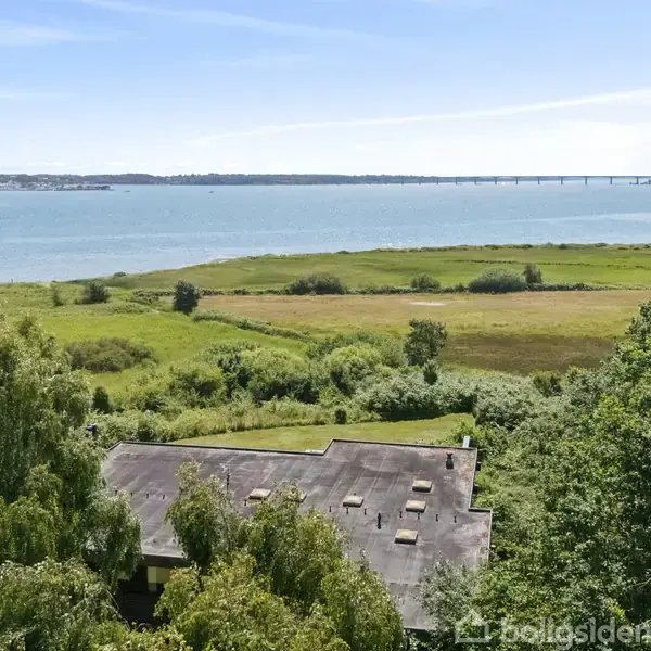 En bygning med fladt tag ses blandt frodig vegetation, omgivet af en bred græsmark. En fjord strækker sig i baggrunden med en langt bro over vandet.