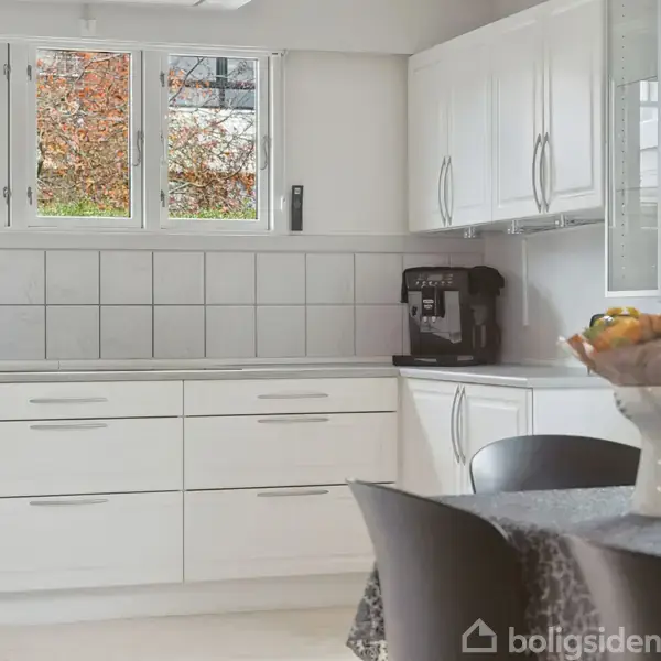 Hvidt køkken med komfur, skabe og en kaffemaskine. Et bord med blomster i en vase står i forgrunden. Udenfor vinduet ses efterårsfarvede træer.