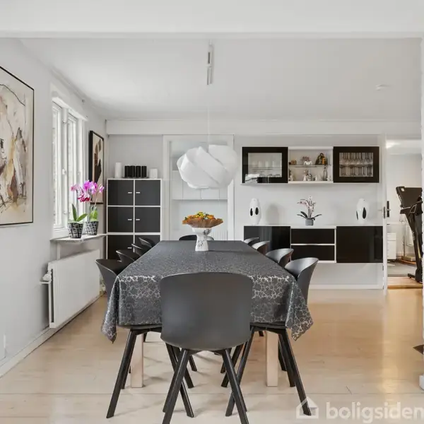 Et spisebord med sort dug og seks sorte stole står i en lys spisestue. Der hænger en hvid lampe over bordet, og blomster pynter i vindueskarmen.