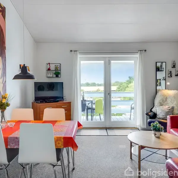 Spisestue med rødt lædersofa, rundt sofabord, farverigt bord med blomster i vase. Glasdøre åbner til terrasse med udsigt til vand og grøn natur. Vægkunst og reoler.