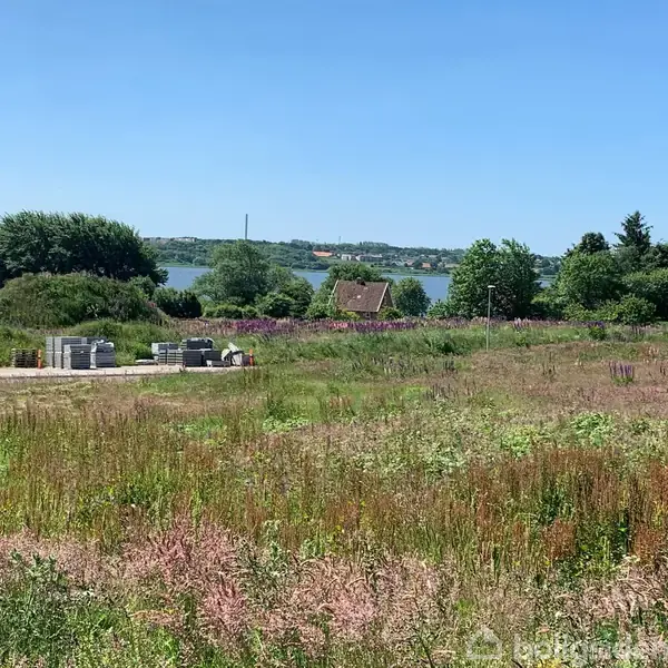 En græseng grænser op til en sø i baggrunden, med træer og buske omkring. I forgrunden ligger en sti med stablede byggematerialer. Himlen er klar og blå.