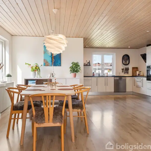 Et spisebord med seks træstole står i et lyst køkkenalrum. Lyse trægulve og et træloft skaber en hyggelig atmosfære. En moderne lampe hænger over bordet.