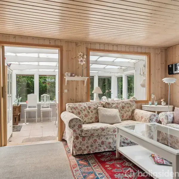 Stue med en blomstret sofa og sofabord. Sofaen står ved siden af et stort vindue med udsigt til en udestue fyldt med naturligt lys. Tæpper dækker gulvet.