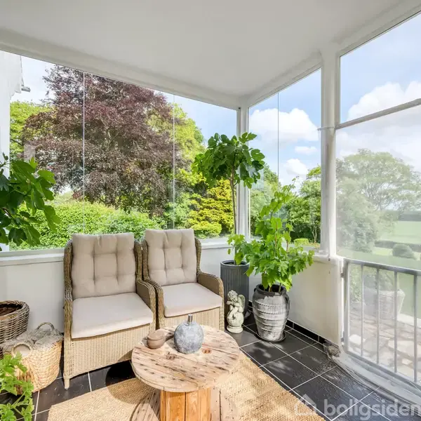 To fletstole står stille på en indendørs terrasse omgivet af glasvægge. Terrassen er dekoreret med grønne planter, et rundt træbord og tæppe. Udsigt over en frodig have.