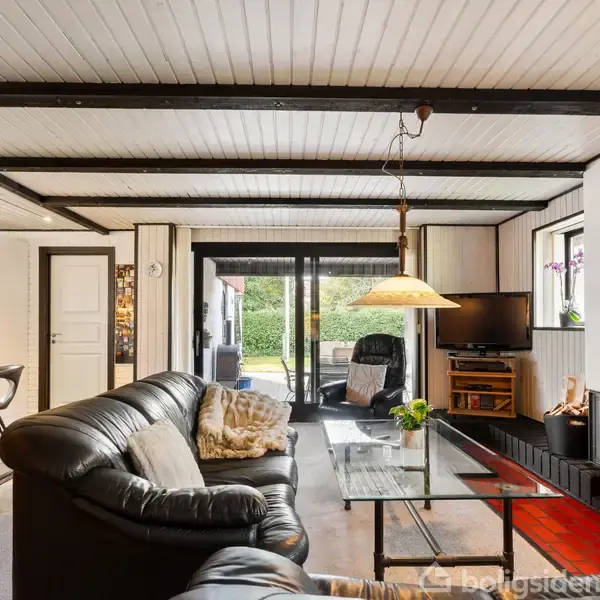 Sort læderhjørnesofa står i en stue med glasbord og tv ved siden af pejs. Store vinduer åbner rummet mod en grøn have.