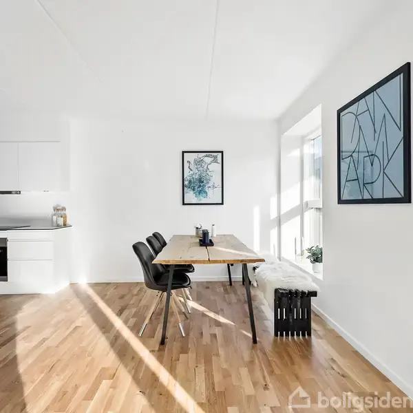 Et lyst køkken-alrum med et træbord og tre sorte stole står på et trægulv. I baggrunden ses et moderne, hvidt køkken. Vægge har kunstværker, herunder en plakat med teksten "DENMARK".