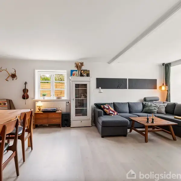 En stue med en stor grå hjørnesofa fyldt med puder, placeret ved vinduer, der lukker lys ind. Ved siden af står et træbord med stole, omgivet af kunst og dekorationer.