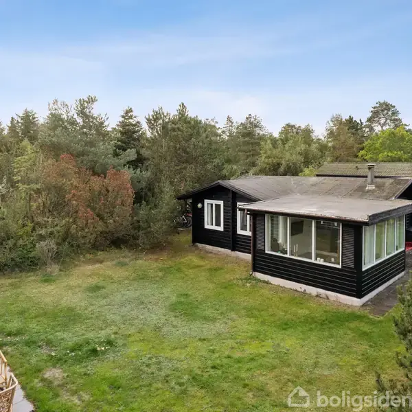 Sort træsommerhus med store vinduer står i en grøn have omgivet af træer. Huset har en veranda og ligger i et fredeligt, skovklædt område.