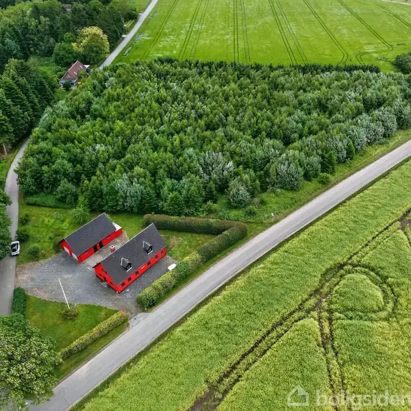 Rødt hus med sort tag står stille omgivet af skove og marker. Veje strækker sig gennem det grønne landskab. Atmosfæren er fredfyldt og landlig.