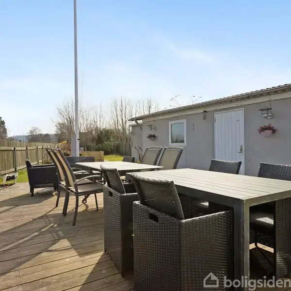Terrassemøbler står stille på et solrigt trædæk ved siden af et gråt hus med vinduer og dør. Omgivet af et plankeværk og spredte potteplanter.