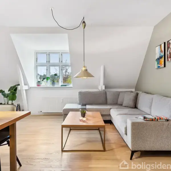 Grå hjørnesofa med puder står i lys stue. Et sofabord med glas på overfladen. Et spisebord med blomster. Vinduet i baggrunden og billeder hænger på væggen.