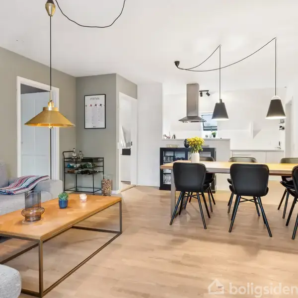 En moderne stue med en grå sofa og et træbord. Der er en spiseplads med seks sorte stole omkring et bord. Loftslamper hænger over bordet, omgivet af lyse vægge og trægulv.