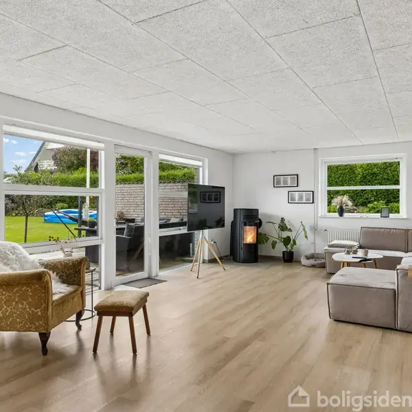 Stue med beige sofa og lænestol ved siden af et sofabord. Store vinduer giver udsigt til en frodig have. En brændeovn og væghylder tilføjer hygge.