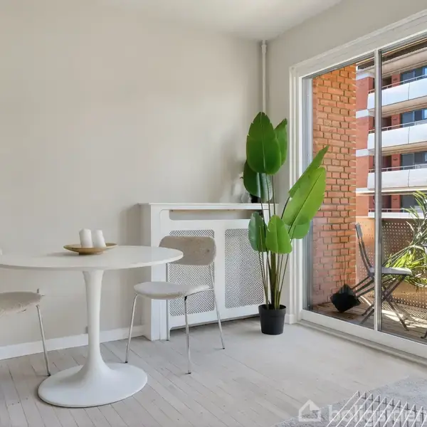 Et rundt, hvidt spisebord står med to stole i et lyst rum. En potteplante er placeret ved siden af bordet. En glasdør åbner ud til en balkon med flere planter.