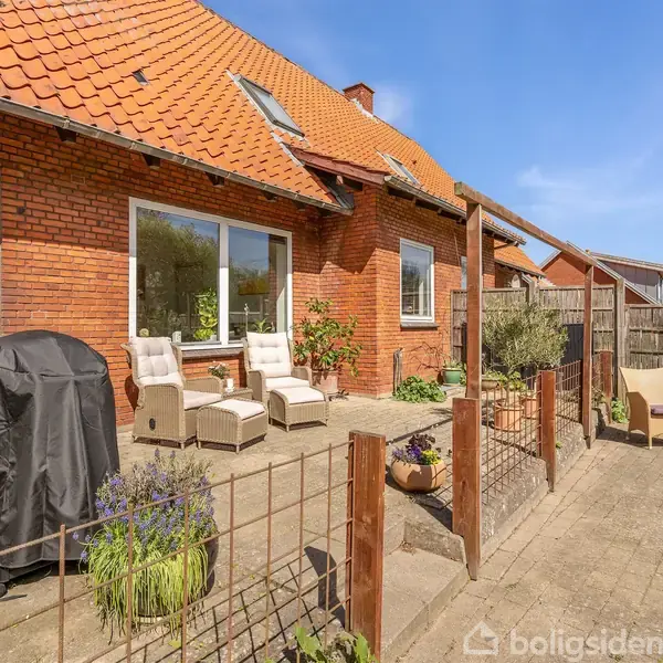 En terrasse foran et rødt murstenshus med tegltag. Terrassemøbler står ved siden af en kulgrill og blomsterkrukker. Træhegn omgiver området under en blå himmel.