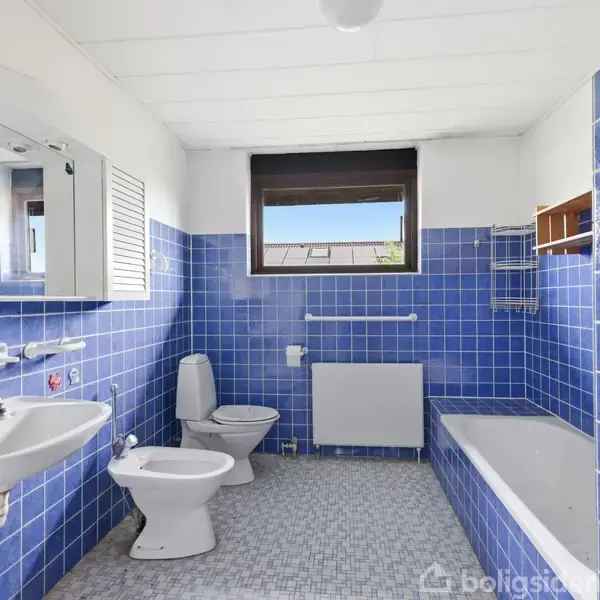 Badeværelse med blå fliser på vægge og gulv. Håndvask, toilet, bidet, og badekar langs væggene. Vindue over en radiator. Spejl og skab er placeret over vasken.