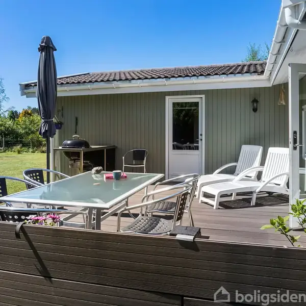 Terrasseområde med glasbord og stole foran et gråmalet sommerhus. Solsenge står ved siden af huset, og en grill er placeret nær døren. Blomster pryder kanten.