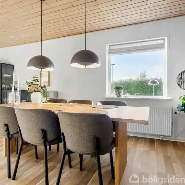 Et spisebord med seks stole står i et lyst rum med træloft. Over bordet hænger tre sorte lamper. En stor vindue giver udsigt til buske udenfor.