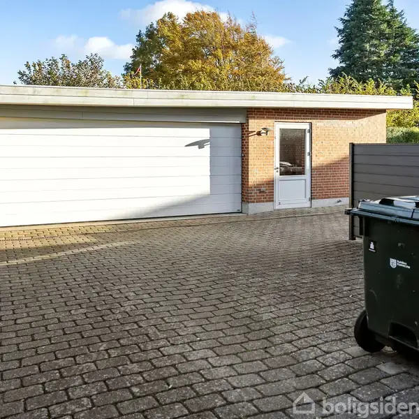 Garagebygning med hvid port og dør på siden set fra en brolagt indkørsel. Skraldespand placeret til højre. Omgivet af grønne hække og træer i efterårsfarver.