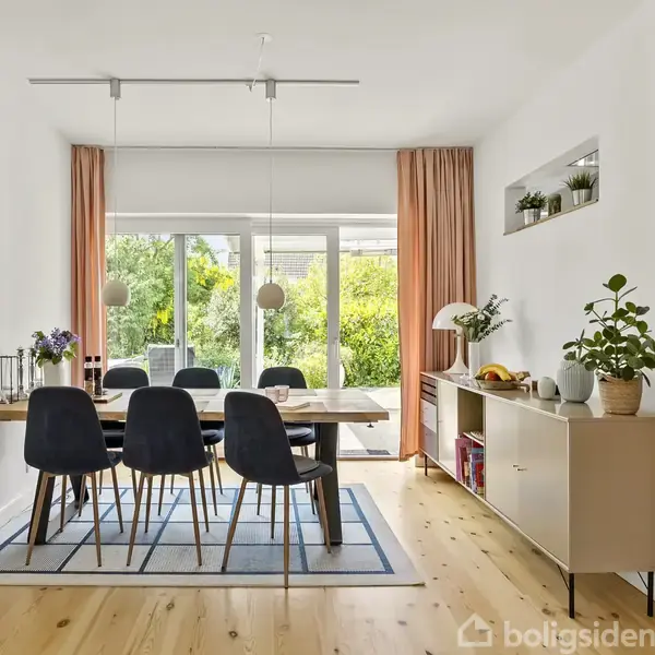 Spisebord med sorte stole står i en lys spisestue. Store vinduer med gardiner viser en have udenfor. Væggen har en murstensdetalje. En skænk står ved siden af med planter og dekorationer.