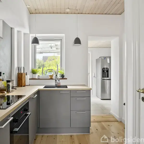Køkken med grå skabe og indbygget ovn. En vask er placeret under et vindue med planter. To lamper hænger fra loftet. Et moderne køleskab ses i baggrunden.