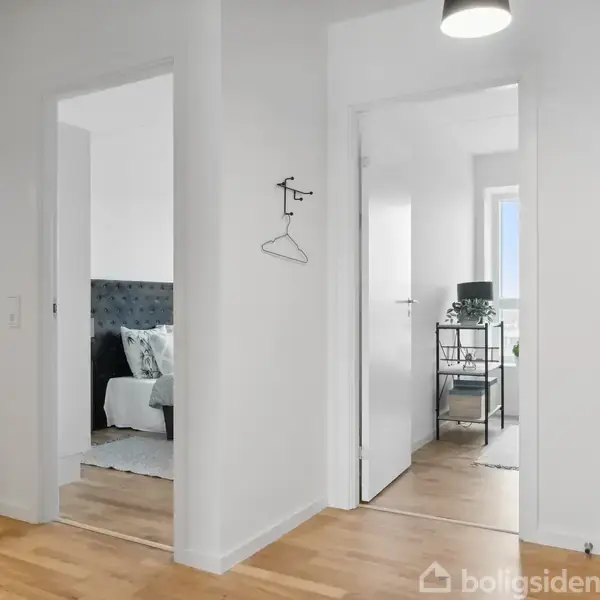 Gang fører til to åbne døre. Venstre dør viser et soveværelse med seng og grå sengegavl. Højre dør afslører en stue med bord og lampe ved vinduet.