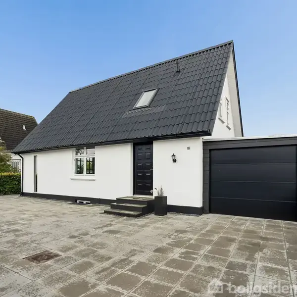Hvidt hus med sort tag står på flisebelagt gårdsplads. Enkelt trappetrin foran hoveddør. Til højre en sort garageport. Omkringliggende have og huse.