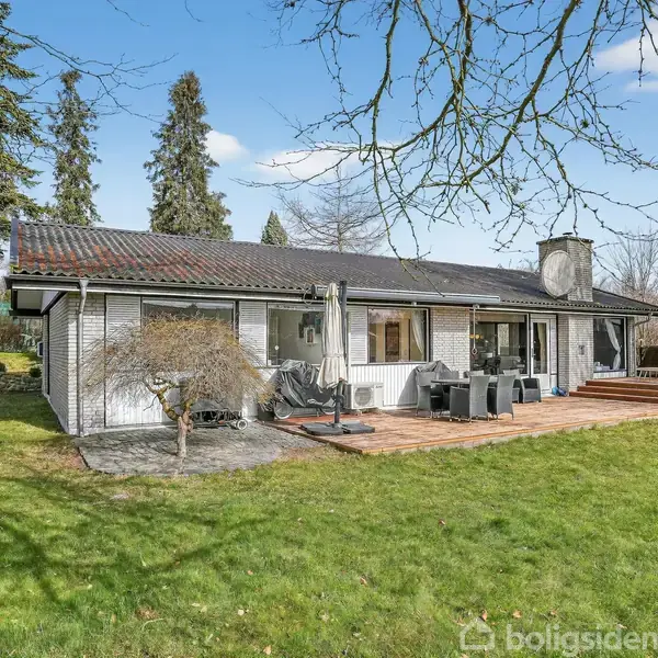 Et moderne hus med et fladt tag står stille omgivet af en stor græsplæne og høje træer. Terrasse med havemøbler ses foran huset.