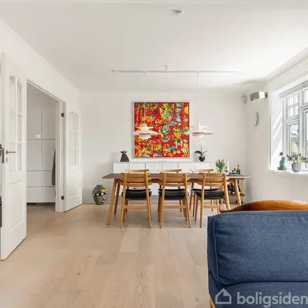 Et spisebord med stole står i en lys stue. Malerier pryder væggene, og en brændeovn er ved døren. Et vindue lader naturligt lys strømme ind fra højre.