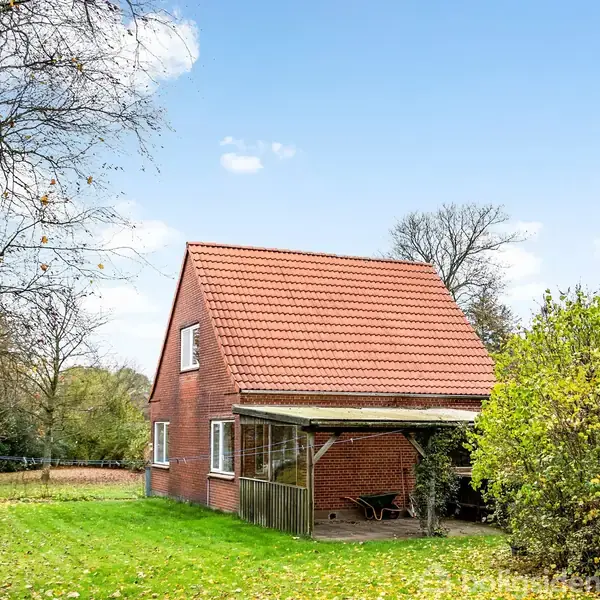Et rødt murstenshus med et rødt tegltag står i en have med grønt græs og træer omkring. Der er en overdækket terrasse med stole tæt på huset under en blå himmel.