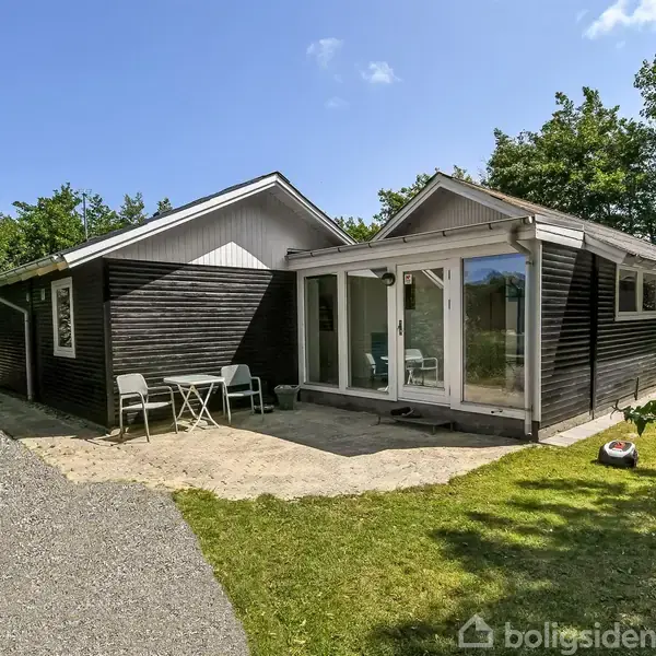 Et sortmalet træhus står stille i et naturskønt område. Et lille plastiksæt bestående af et bord og to stole står ved indgangen. Græsplæne og buske omringer huset.